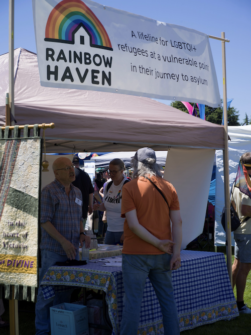 Brian speaking to a visitor at one side of the booth while another visitor inspects the literature on display. The banner on the left belongs to our sponsor the Anglican Church of St. John the Divine Victria.